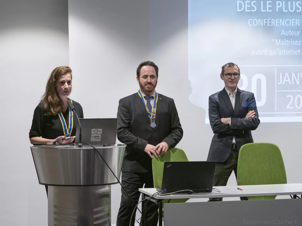 Trois personnes se tiennent à l'avant d'une salle de conférence. Une femme parle de l'éducation numérique sur un podium, tandis que deux hommes se tiennent à proximité. Un ordinateur portable et un projecteur sont posés sur la table, affichant une présentation en arrière-plan. - Rotary Club Paris Agora