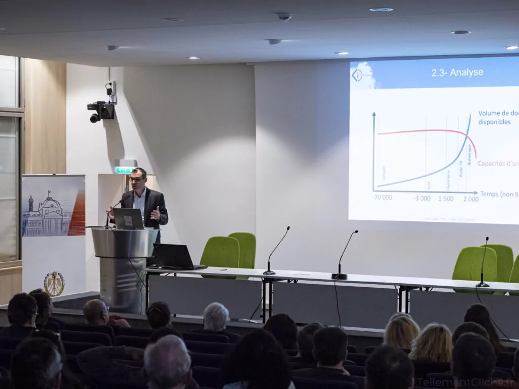 Un homme en costume est debout sur un podium et fait une présentation sur l'éducation numérique à un public. Derrière lui, une diapositive affichant un graphique et un texte est projetée sur un écran, tandis que plusieurs chaises vides se trouvent sur la scène. - Rotary Club Paris Agora