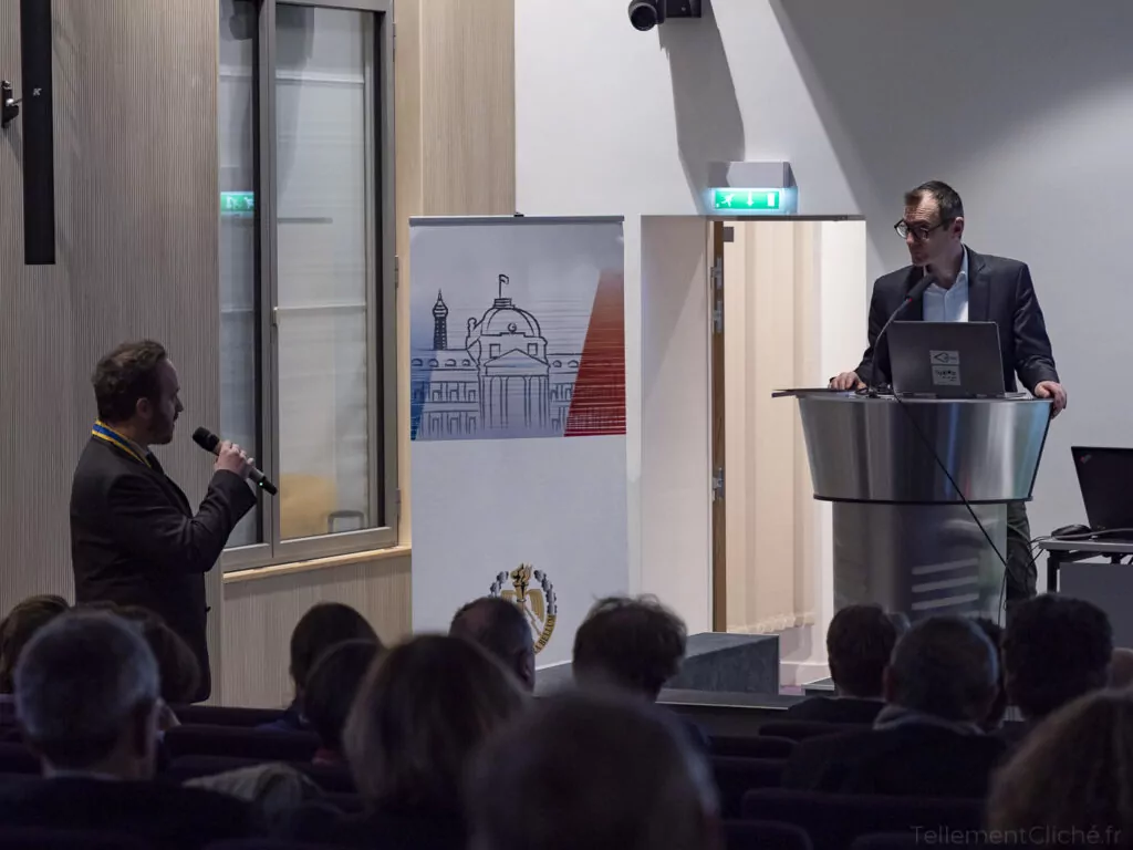 Un homme se tient sur un podium et s'adresse à un auditoire au sujet de l'éducation numérique, tandis qu'un autre homme muni d'un microphone se tient sur le côté. Une banderole représentant un bâtiment et des armoiries est affichée entre eux. - Rotary Club Paris Agora