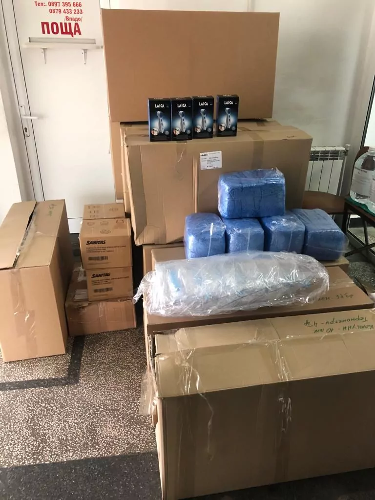 Plusieurs grandes boîtes en carton, des paquets emballés en bleu et des rouleaux emballés en plastique sont empilés dans une pièce carrelée de l'hôpital de Zlatograd. Un panneau avec du texte est visible au-dessus de la porte, marquant ce don de matériel de Sofia International. - Rotary Club Paris Agora