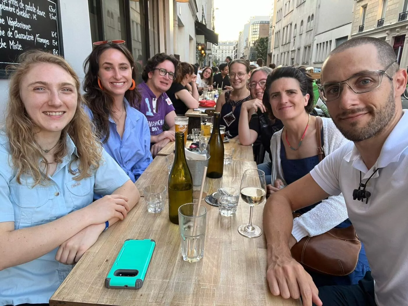 Sept personnes sont assises autour d'une table en bois dans un restaurant de la ville et sourient à l'appareil photo. Des boissons, des bouteilles, des verres et un téléphone sarcelle avec un étui noir sont posés sur la table. Ils discutent de Bourses d'études tandis que des bâtiments et d'autres convives se trouvent à l'arrière-plan. - Rotary Club Paris Agora