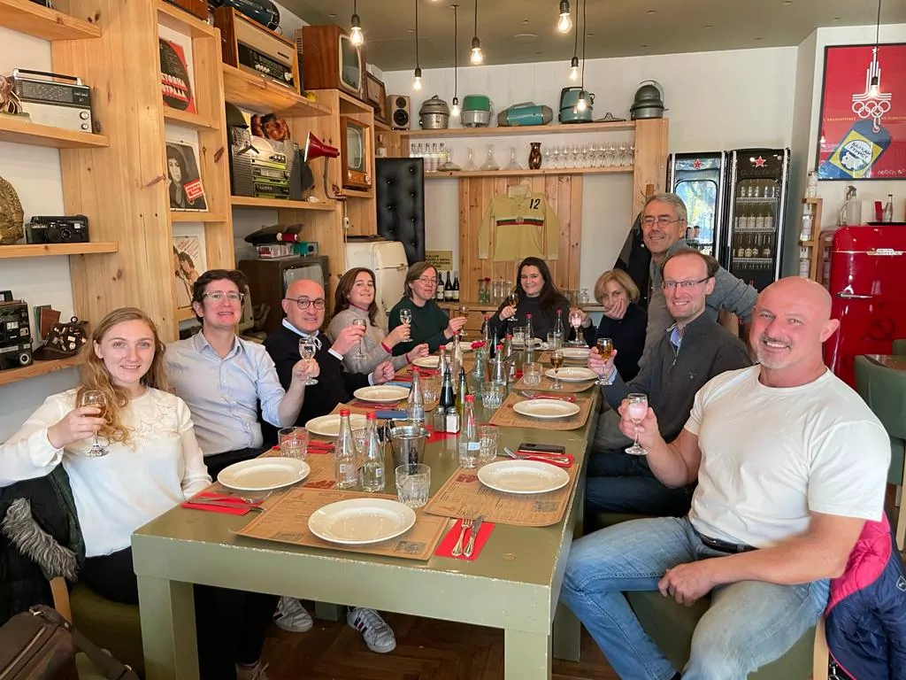 Nine people are seated around a rectangular table, raising their glasses and smiling in a casual restaurant where wooden shelves display antique objects - a scene celebrating the friendship between Jumelage and Sofia International. - Rotary Club Paris Agora