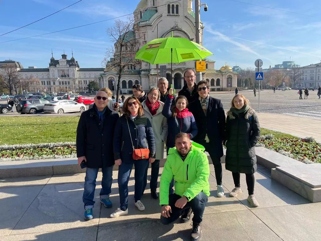 A group of ten people, some standing and one kneeling in front, pose for an outdoor photo on a sunny day during a Twinning event. One person holds a bright green umbrella, with historic buildings and parked cars in the background. - Rotary Club Paris Agora