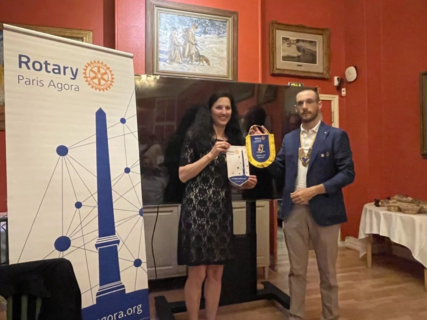 A woman and a man stand indoors next to a banner that reads "Rotary Paris Agora," celebrating the twinning with Sofia International. The woman holds a small banner, and the man wears a medallion. Artwork is visible on the red walls in the background. - Rotary Club Paris Agora