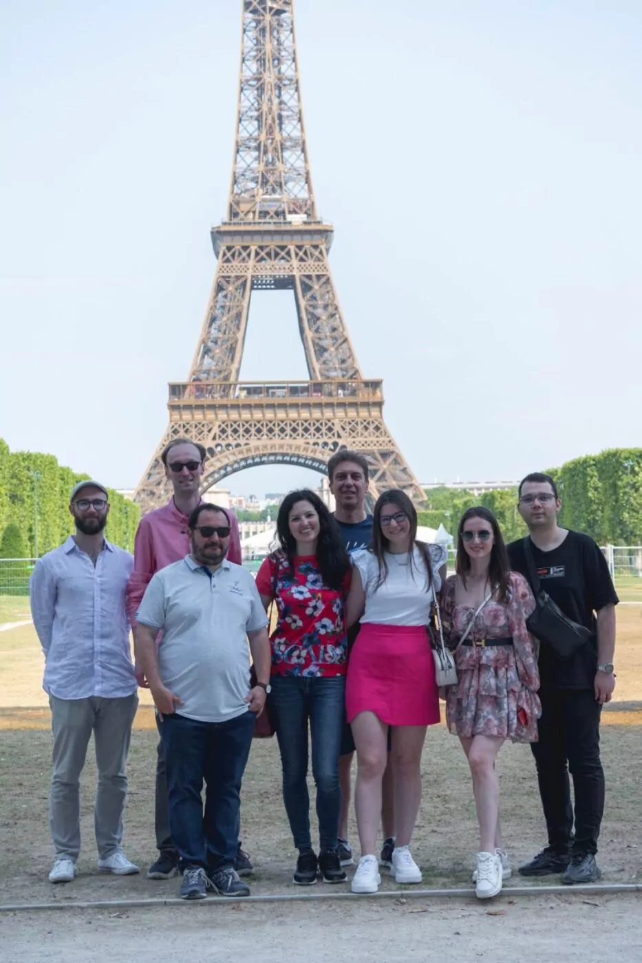 A group of eight people poses together for a photo in front of the Eiffel Tower on a sunny day, celebrating their twinning with Sofia International, surrounded by green trees and a clear sky in the background. - Rotary Club Paris Agora