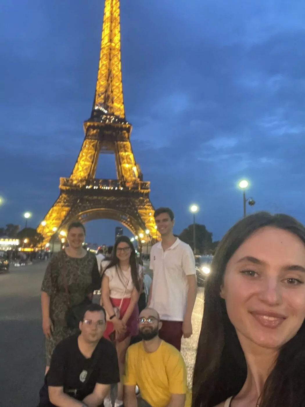 A group of six people poses in front of the illuminated Eiffel Tower at dusk, celebrating a Twinning event. The sky is cloudy, lampposts shine behind them, and a person from Sofia International stands in the foreground, smiling at the camera. - Rotary Club Paris Agora