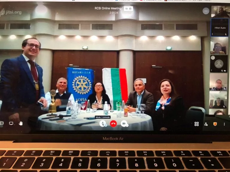 A group of five people is sitting at a round table with documents, water bottles, and a flag in a conference room. A Rotary Club banner and a Sofia International twinning event are visible on a laptop screen. A man is standing on the left. - Rotary Club Paris Agora