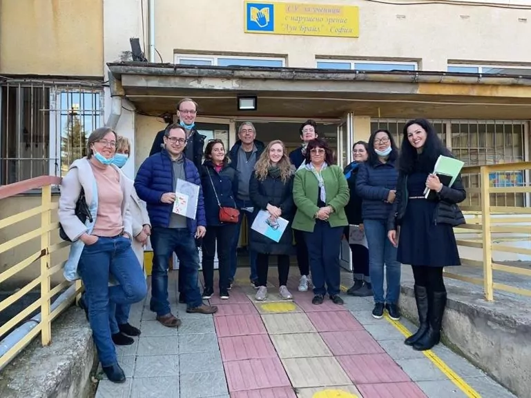 A group of twelve adults, some of whom are holding papers and files, stand outside a building with a yellow sign and railing. Most of them are smiling and dressed casually. They are gathered for the Sofia International Twin Event. The building has large windows and a ramp. - Rotary Club Paris Agora