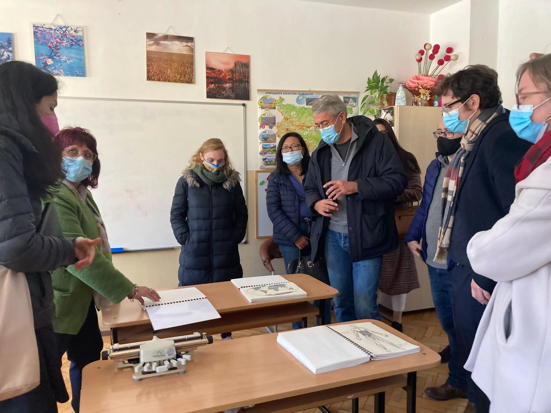 A group of adults wearing masks are gathered around desks in a classroom, looking at open binders and tactile diagrams during a Matching activity, while one woman gestures toward the materials. A whiteboard and posters are mounted on the walls. - Rotary Club Paris Agora