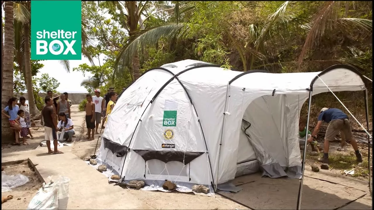 Une grande tente d'abri d'urgence blanche portant les logos de ShelterBox et du Rotary est installée en plein air sur un chemin en béton, entourée d'arbres et de personnes se tenant ou travaillant à proximité dans un environnement tropical. - Rotary Club Paris Agora