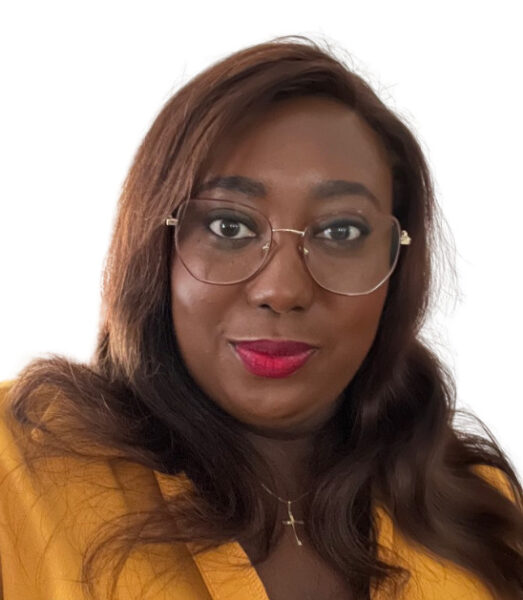 N'Doumi-Daniel, une femme aux longs cheveux bruns et aux lunettes, portant un haut jaune moutarde et un collier doré, pose sur un fond blanc uni. Elle porte un rouge à lèvres rose vif et regarde directement l'appareil photo. - Rotary Club Paris Agora
