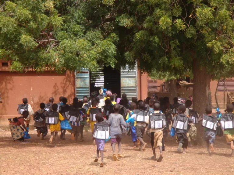 A group of schoolchildren with backpacks running towards the entrance of a building in Burkina Faso, partially shaded by a large tree, with dry ground, scattered leaves, and a few bicycles parked nearby. - Rotary Club Paris Agora