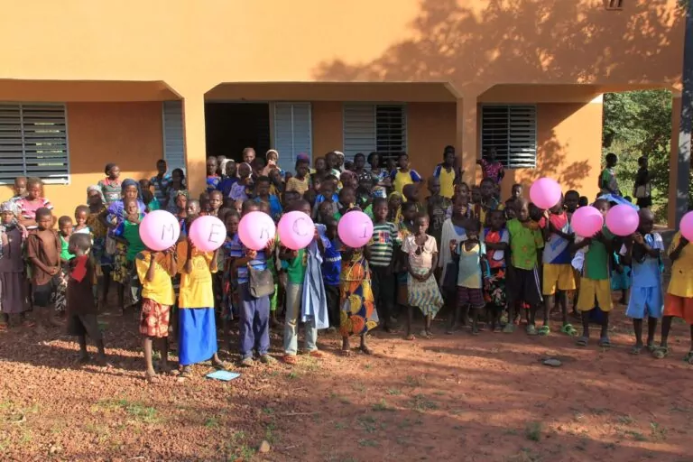 A large group of schoolchildren stands in front of a beige building in Burkina Faso. A few children in the front hold pink balloons with "THANK YOU" written on them. The red-brown dirt ground and sunlight cast strong shadows on this joyful scene. - Rotary Club Paris Agora