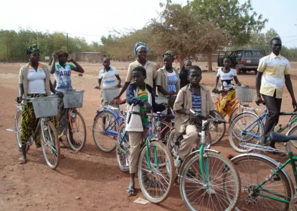 Un groupe d'écoliers du Burkina-Fasso, dont certains portent des vêtements à motifs et des foulards, se tient à l'extérieur avec des vélos sur un chemin de terre. Des arbres, un véhicule garé et des bâtiments sont visibles à l'arrière-plan. - Rotary Club Paris Agora
