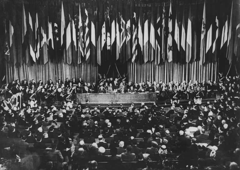 A large formal assembly is seated facing a stage, where officials speak at a long table, discussing the Rotary's role in creating UNESCO, with national flags behind them. The audience fills the hall while musicians are seated on the sides of the stage. - Rotary Club Paris Agora