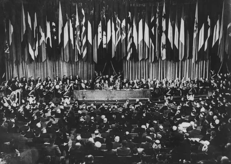 A large formal assembly is seated facing a stage, where officials speak at a long table, discussing the Rotary's role in creating UNESCO, with national flags behind them. The audience fills the hall while musicians are seated on the sides of the stage. - Rotary Club Paris Agora