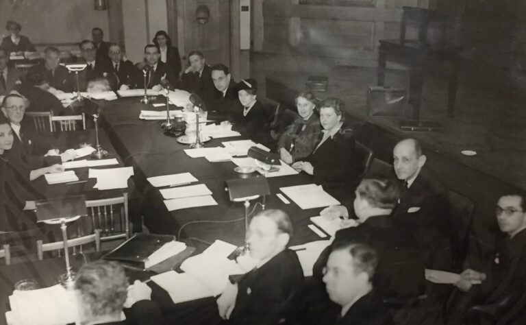 A group of men and women in formal attire are seated around a large table covered with papers and lamps in a meeting room, likely attending a Rotary conference on the role in the creation of UNESCO. - Rotary Club Paris Agora