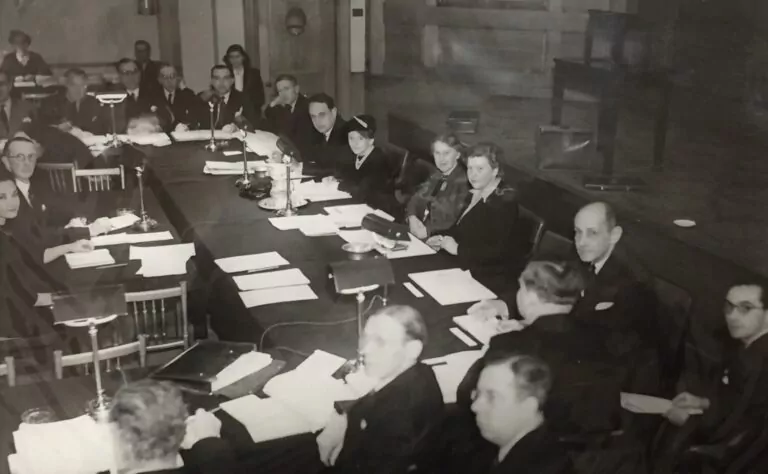 A group of men and women in formal attire are seated around a large table covered with papers and lamps in a meeting room, likely attending a Rotary conference on the role in the creation of UNESCO. - Rotary Club Paris Agora