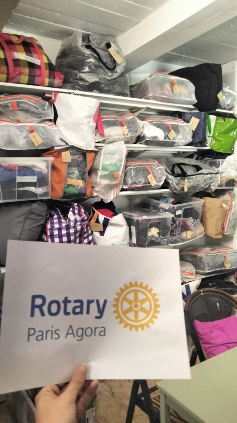 Une main tient un panneau indiquant "Rotary Paris Agora" devant des étagères remplies de sacs et de colis organisés provenant d'une collection de textiles, chacun étiqueté et stocké dans des conteneurs transparents dans une salle de stockage. - Rotary Club Paris Agora