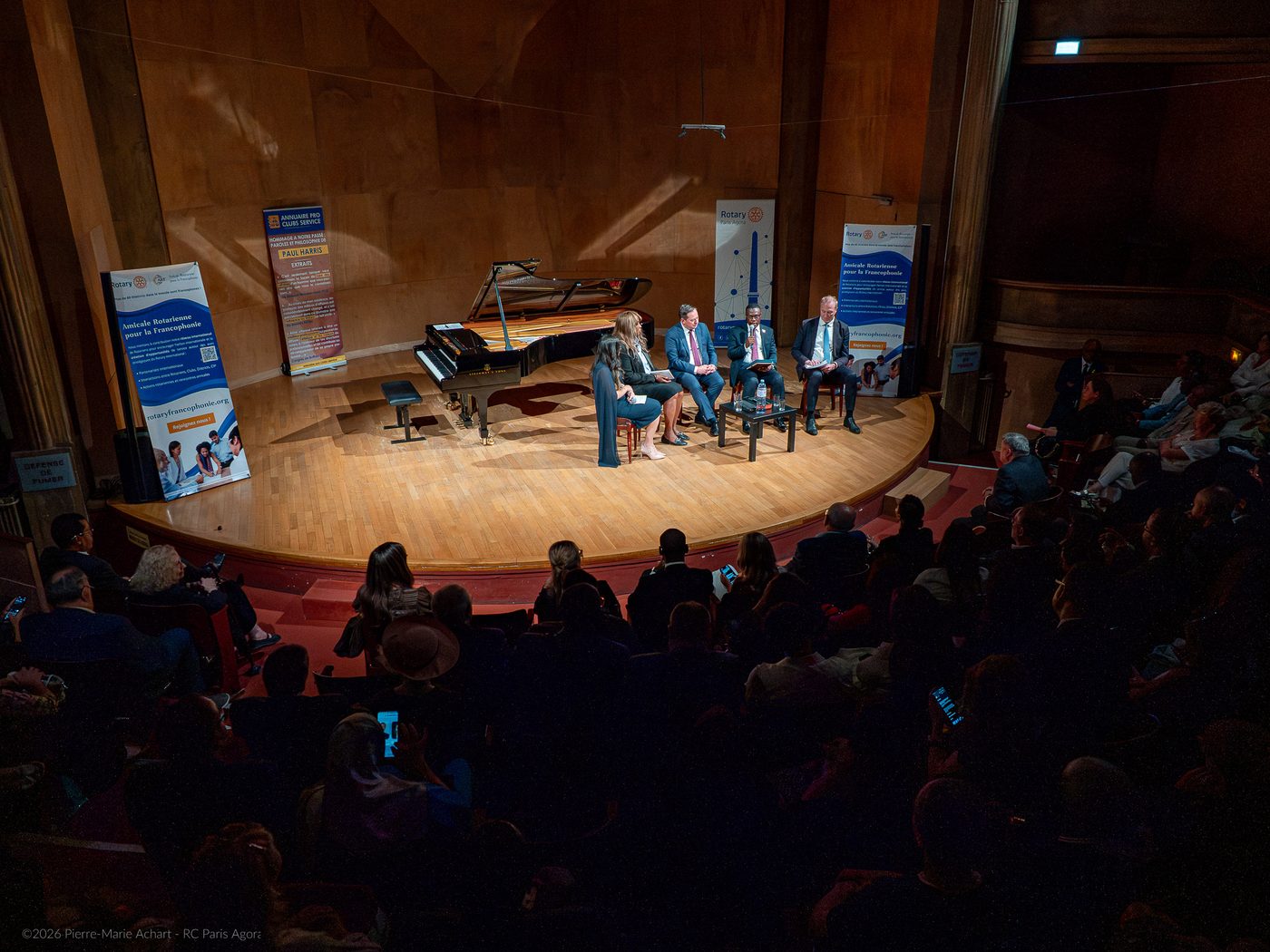 Quatre personnes sont assises sur une scène lors d'une table ronde organisée dans le cadre du Symposium pour la Paix, dans une salle de concert, avec le public visible au premier plan. - Rotary Club Paris Agora