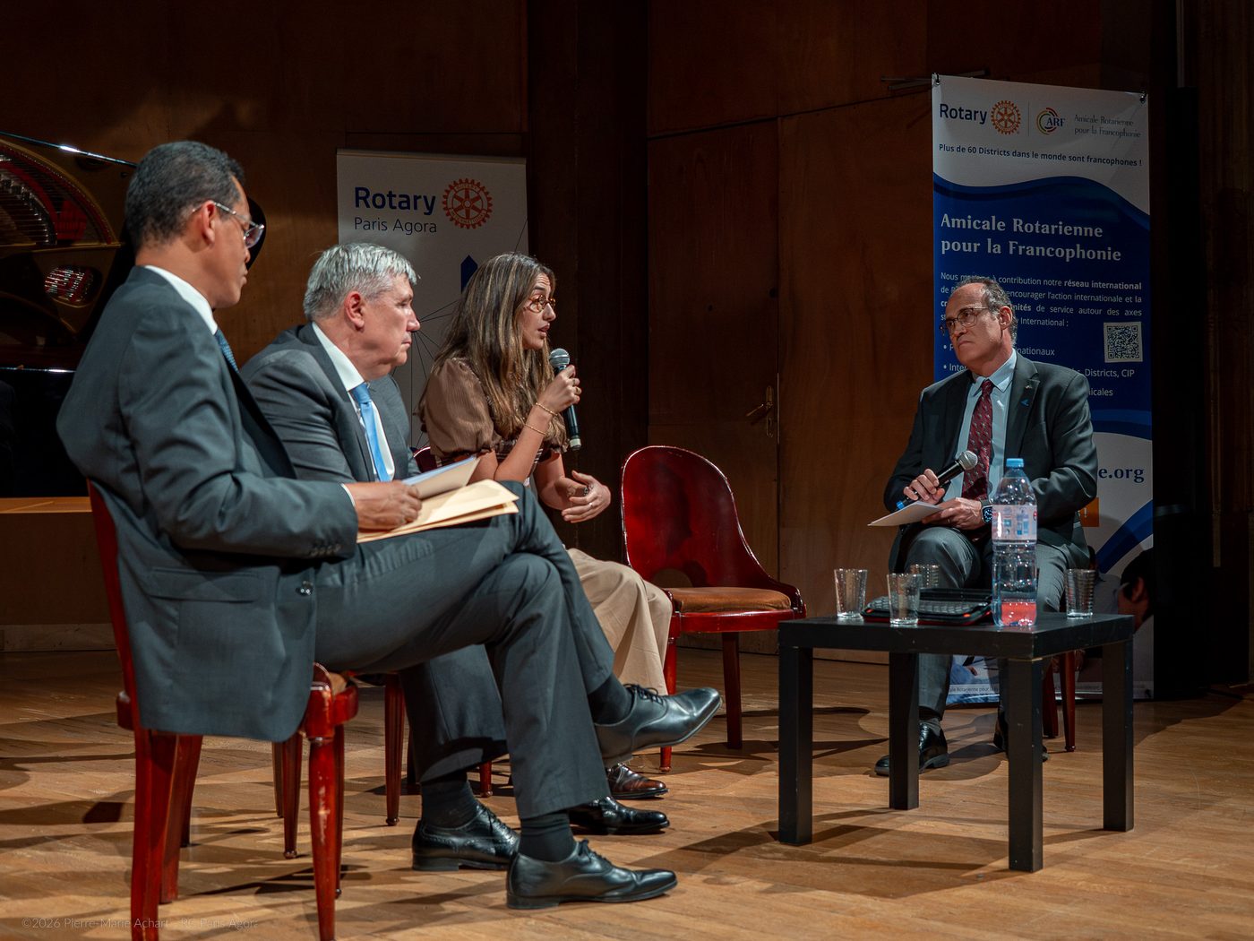 Quatre personnes sont assises sur une scène lors d'une table ronde organisée dans le cadre du Symposium pour la Paix ; une femme prend la parole au micro. - Rotary Club Paris Agora
